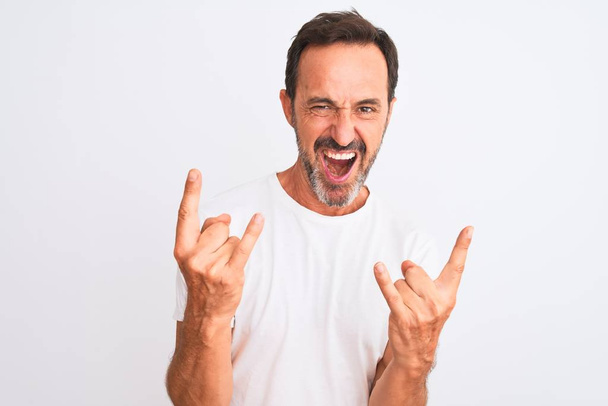 Ein gutaussehender Mann mittleren Alters in lässigem T-Shirt steht vor isoliertem weißen Hintergrund und schreit mit verrücktem Gesichtsausdruck Rocksymbole mit erhobenen Händen. Musikstar. Schweres Konzept. - Foto, Bild