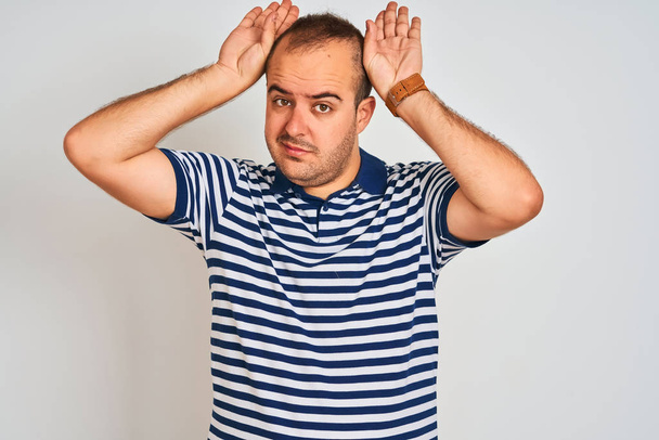 Young man wearing casual striped polo standing over isolated white background Doing bunny ears gesture with hands palms looking cynical and skeptical. Easter rabbit concept. - Fotoğraf, Görsel