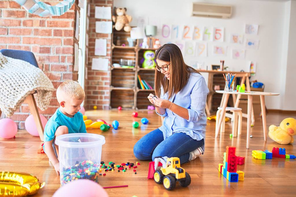 Young caucasian child playing at playschool with teacher. Mother and son at playroom bulding a tower with toy blocks - Photo, Image