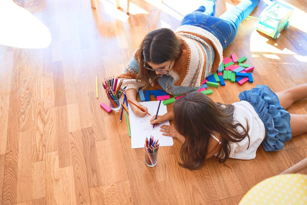Schöne Lehrerin und Kleinkind zeichnen im Kindergarten mit Bleistift und Papier um jede Menge Spielzeug herum - Foto, Bild