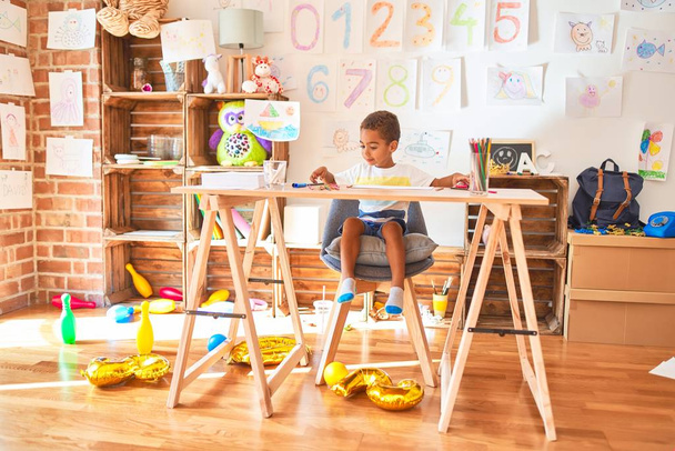 Hermoso niño afroamericano sentado dibujo usando papel y lápices en el escritorio en el jardín de infantes
 - Foto, imagen