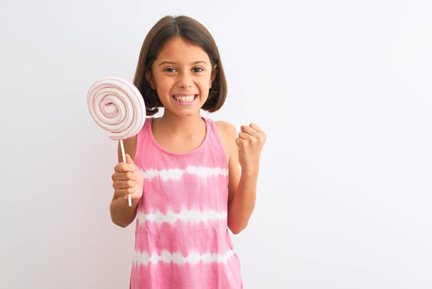 Beautiful child girl eating sweet lollipop standing over isolated white background screaming proud and celebrating victory and success very excited, cheering emotion - Photo, Image