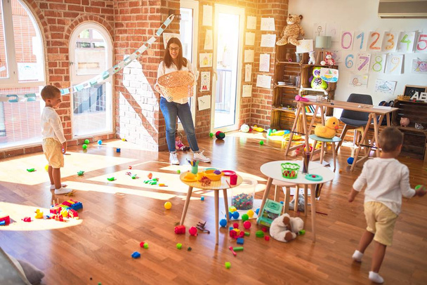 Красивий вчитель і малюки грають в баскетбол за допомогою кульок і плетеного кошика навколо великої кількості іграшок в дитячому саду
 - Фото, зображення