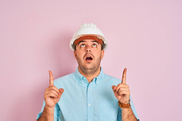 Young architect man wearing security helmet standing over isolated pink background amazed and surprised looking up and pointing with fingers and raised arms. - Fotoğraf, Görsel