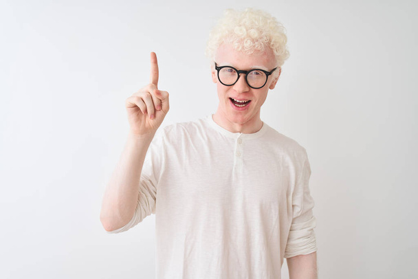 Mladý albín blonďatý muž v tričku a brýlích, stojící nad izolovaným bílým pozadím a ukazující prstem nahoru s úspěšným nápadem. Utekl a byl šťastný. Číslo jedna. - Fotografie, Obrázek
