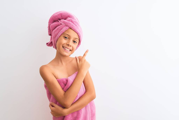 Beautiful child girl wearing shower towel after bath standing over isolated white background cheerful with a smile of face pointing with hand and finger up to the side with happy and natural expression on face - Foto, afbeelding