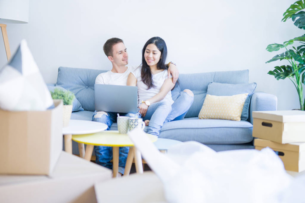 Young beautiful couple sitting on the sofa using laptop at new home around cardboard boxes - Foto, Imagen