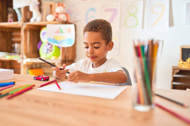 Красивый африканский американец сидит рисовать бумагой и карандашами на столе в детском саду
 - Фото, изображение