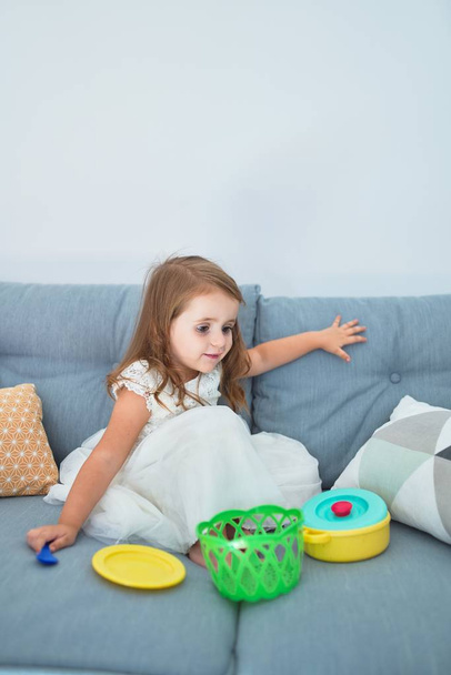 Urocze blond maluch siedzi na kanapie gotowanie z jedzeniem i sztućce zabawki w domu - Zdjęcie, obraz