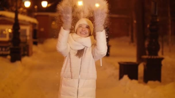 Młoda szczęśliwa dziewczyna rzuca śniegiem na nocną ulicę miasta - Materiał filmowy, wideo