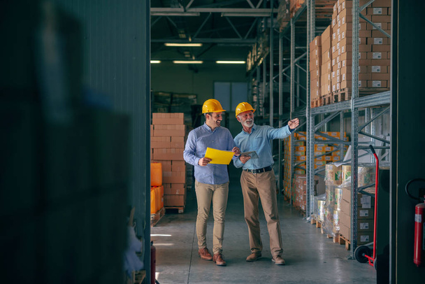 CEO going around warehouse with supervisor and talking analyzing sale statistics. Younger man holding folder with data while older one holding tablet and pointing at boxes. Both having yellow helmets. - Photo, Image