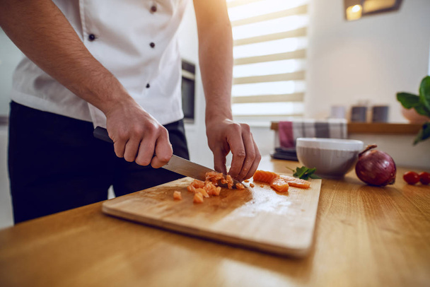 Primer plano del digno chef caucásico dedicado en uniforme cortando salmón en trozos pequeños. Interior de la cocina doméstica
. - Foto, imagen