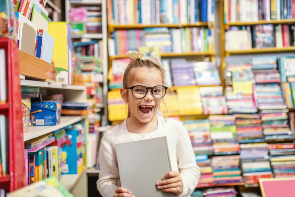 Ενθουσιασμένη καυκάσιος χαριτωμένο μικρό ξανθό κορίτσι με γυαλιά ηλίου και αλογοουρά στέκεται στο βιβλιοπωλείο και κρατώντας το βιβλίο που έψαχνε. - Φωτογραφία, εικόνα