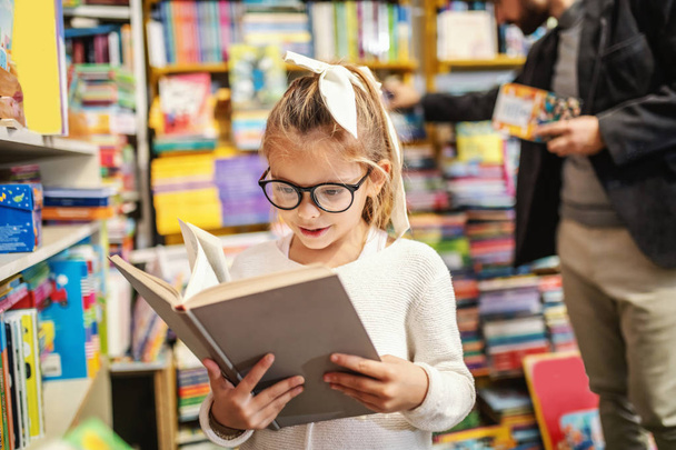 Adorable curious caucasian little girl with eyeglasses standing in bookstore and reading interesting book. All around are books on shelves. - 写真・画像