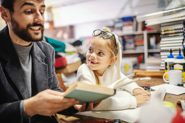 Αξιολάτρευτο μικρό καυκάσιο ξανθό κορίτσι χωρίς δύο δόντια να διαβάζει βιβλίο στον στοργικό πατέρα του στο βιβλιοπωλείο.. - Φωτογραφία, εικόνα