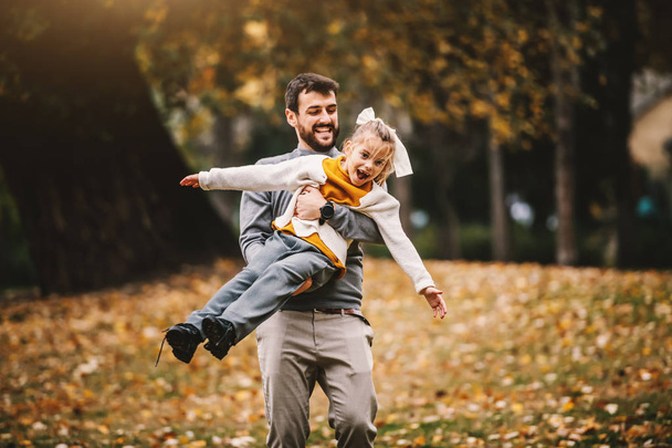 Zabawny śmiejący się biały brodaty ojciec bawiący się ze swoją ukochaną córką w parku. Czas jesieni. - Zdjęcie, obraz