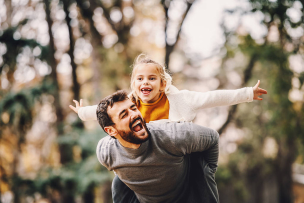 Playful handsome bearded Caucasian father having piggyback with his adorable daughter. Autumn in park. - Foto, Imagem