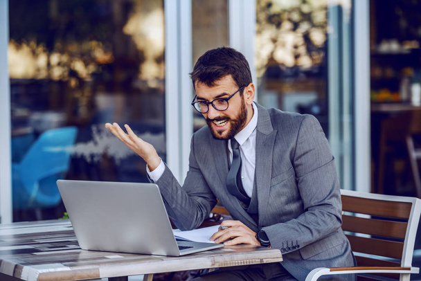 Jovem atraente barbudo sorridente caucasiano empresário escrever tarefas na agenda e olhando para laptop. Café exterior
. - Foto, Imagem