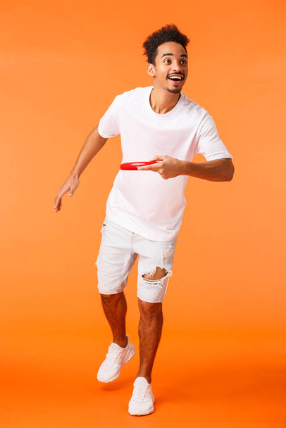 Full-length vertical shot happy, charismatic african-american sporty guy like active games outdoors, throwing frisbee at friend and smiling, enjoying beautiful sunny summer day, orange background - Valokuva, kuva