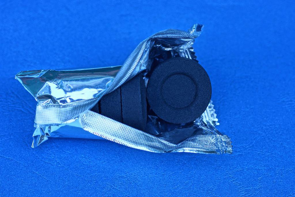 un conjunto de carbones redondos negros para una cachimba en un paquete de lámina abierta gris se encuentra en una mesa azul
 - Foto, imagen