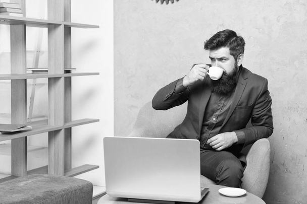 Drinking coffee for breakfast. Businessman drinking coffee at workplace. Bearded man holding drinking cup at laptop. Enjoy drinking tea or other hot beverage - Фото, изображение