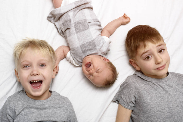 Dois irmãos mais velhos e o bebé mais novo numa cama branca. Infância feliz, família grande. Vista superior
 - Foto, Imagem