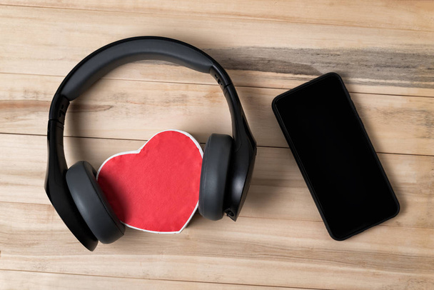 Πλήρες μέγεθος ασύρματα ακουστικά τράβηξε πάνω από μικρό κόκκινο καρδιά σχήμα κουτί και smartphone σε ανοιχτό καφέ ξύλινο τραπέζι. Ακριβώς από πάνω - Φωτογραφία, εικόνα