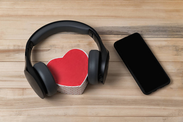 Full-size wireless headphones pulled over small red heart-shaped box and smartphone on light brown wooden table - Photo, Image