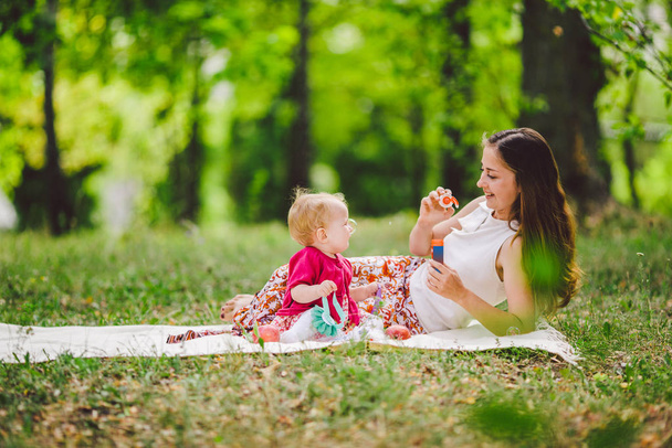 Smiling woman play and sit on green grass in park, rest and hug hold soap bubble blower with little cute child baby girl. Happy mother and daughter in the park. Family outdoor recreation - Photo, Image