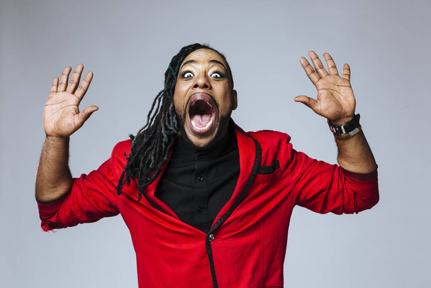 Studio portrait of a man wearing a red jacket suit and dreadlocks with both hands up and screaming - Photo, Image