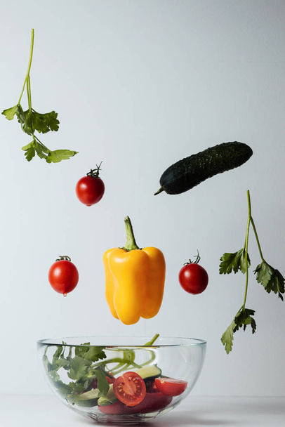 Здоровый салат с летящими растительными ингредиентами на белом фоне. Помидоры черри, желтый перец, огурец, петрушка
 - Фото, изображение