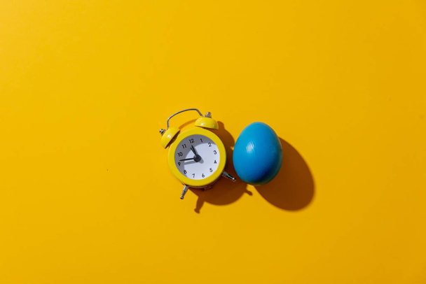Huevo azul de Pascua y despertador sobre fondo amarillo
 - Foto, imagen