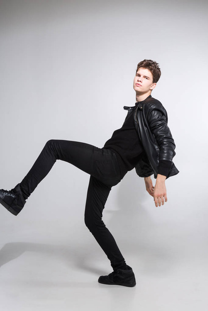 Caucasian young guy posing in studio, full-length portrait of a model in black clothes. Handsome teenager in a black jacket, jeans and sneakers on a gray background. Man in a leather jacket - Fotografie, Obrázek