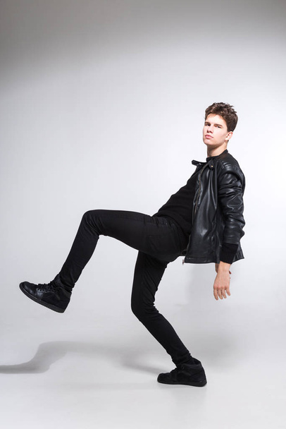 full length portrait of young fit man in dark cloths on the white background. Young Male Fashion Model Posing In Casual Outfit. Attractive young fashion model wearing black in leather jacket - Zdjęcie, obraz