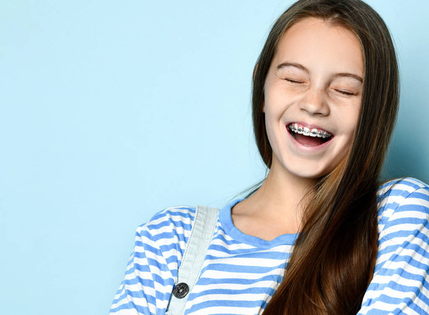 Cheerful beautiful young woman with dark hair and braces on teeth laughing isolated on a blue background. - 写真・画像