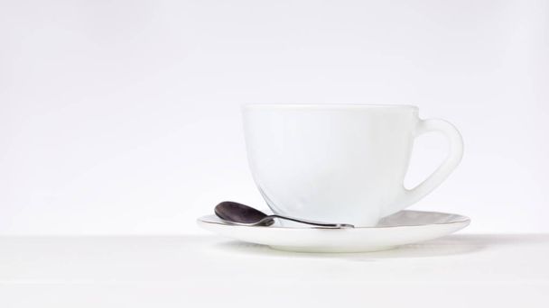 Una taza de té o café blanco y una cuchara de metal sobre una mesa blanca sobre un fondo gris
. - Foto, Imagen