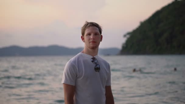 Porträt eines jungen, gut aussehenden, reisenden Mannes, der in die Kamera blickt, mit einer Szene des tropischen Sonnenuntergangs hinter der Insel - Filmmaterial, Video