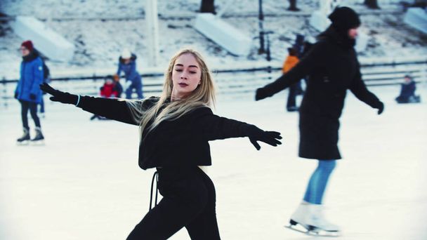 Een jonge blonde aantrekkelijke vrouw professionele kunstschaatser schaatsen op de buitenlucht ijsbaan rond mensen - Foto, afbeelding