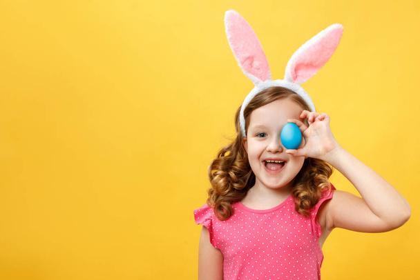 Szczęśliwe dziecko w króliczych uszach trzyma wielkanocne niebieskie jajko. Portret dziewczynki na żółtym tle - Zdjęcie, obraz