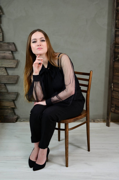 Full-length photo of a pretty brunette girl with excellent make-up in dark clothes sitting on a stand on a gray background in a luxury vintage interior. The concept of a glamorous portrait. - Valokuva, kuva