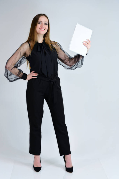 Concept portrait for a banner. Full-length photo of a pretty smiling brunette girl with a folder in her hands with excellent makeup in dark clothes is standing on a white background. - Photo, image