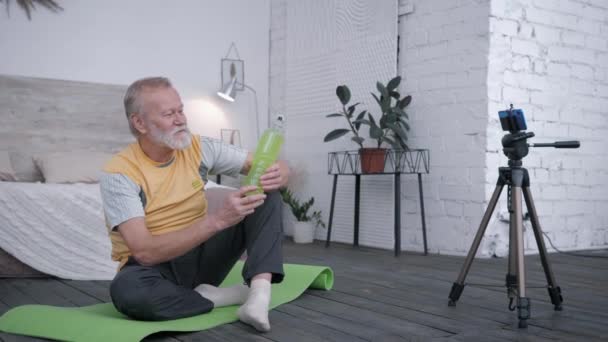 oude blogger, ouder wordende man Influencers houden van sport neemt een drankje review op smartphone voor vlog aanbevelen van water aan abonnees tijdens het zitten op yoga mat in creatievelingen interieur - Video