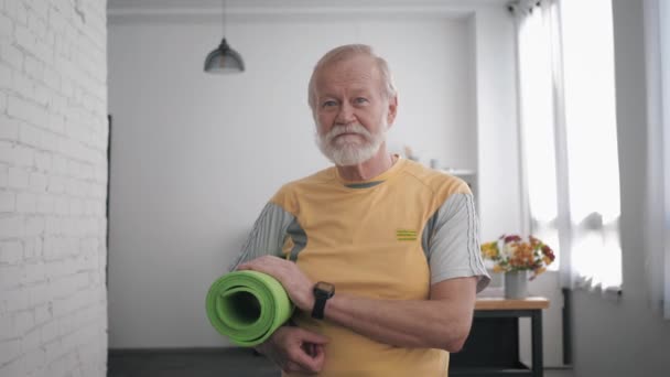 Portret van een knappe oudere man met een yogamat in zijn handen en een slim horloge na het sporten om zijn gezondheid staande te houden in de ruimte tegen een prachtig interieur - Video