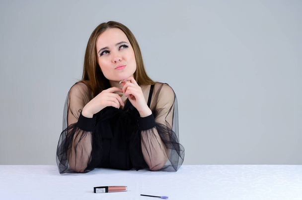 Woman is smiling beautifully at the camera, shows emotions. Photo of a cute brunette with excellent makeup in dark clothes sitting at a white table on a white background. - Foto, Bild