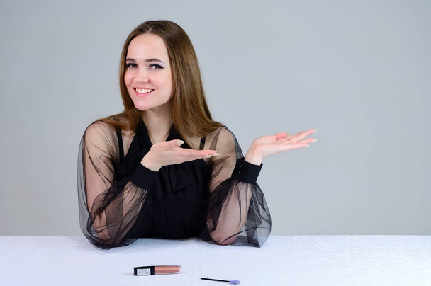Photo of a cute brunette with excellent makeup in dark clothes sitting at a white table on a white background. He smiles beautifully at the camera, shows emotions. - Valokuva, kuva