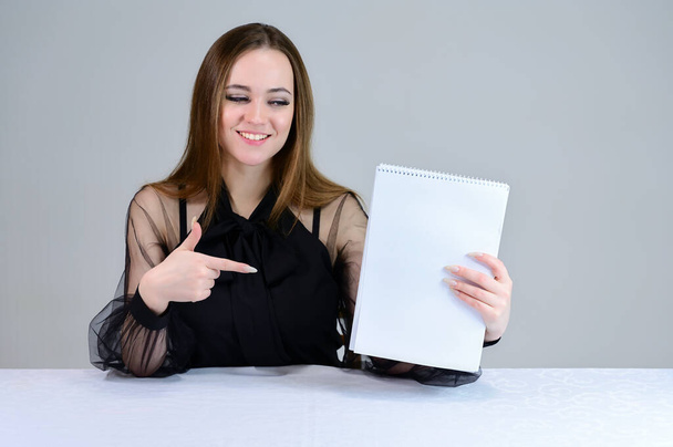 Woman with a folder in her hands. She smiles beautifully at the camera, shows with her hands. Photo of a cute brunette with great makeup in dark clothes sitting at a table on a white background. - Valokuva, kuva