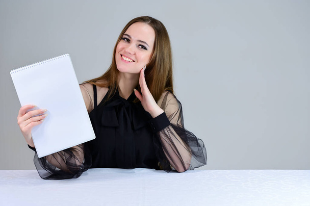 Woman with a folder in her hands. She smiles beautifully at the camera, shows with her hands. Photo of a cute brunette with great makeup in dark clothes sitting at a table on a white background. - Фото, зображення