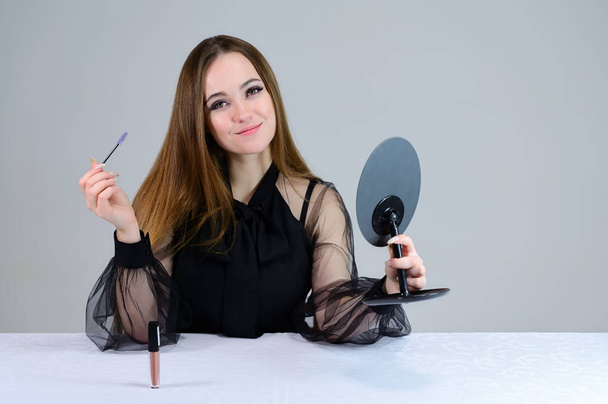 A woman does makeup herself in front of a mirror. Photo of a cute brunette with great makeup in dark clothes sitting at a table on a white background. - Fotografie, Obrázek