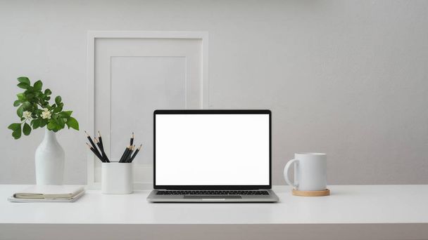 Captura recortada de espacio de trabajo con computadora portátil de pantalla en blanco, marco, lápices, taza de café y jarrón en la mesa blanca con pared blanca
  - Foto, imagen