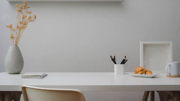 Close-up zicht op werkruimte met kopieerruimte, decoraties, briefpapier, koffiebeker en croissant op witte tafel met witte wand  - Foto, afbeelding